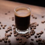 A close-up of a rich espresso in a glass surrounded by coffee beans on a wooden table.